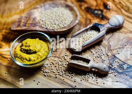 Moutarde blanche (Sinapis alba, Brassica alba), graines de moutarde dans un bol en bois et des cuillères et moutarde prête à l'emploi dans un bol en verre Banque D'Images