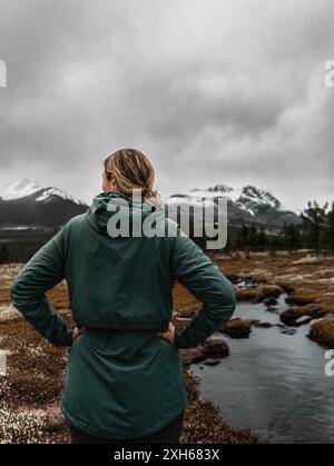 Une femme randonneuse itinérante se tient les mains sur les hanches, regardant un sommet de montagne enneigé et une rivière sinueuse dans la région nordique de Laponie Banque D'Images