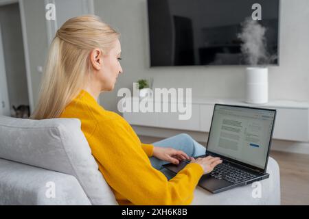 Femme blonde dans les types de chandails sur ordinateur portable dans le salon bien éclairé, créant un espace de travail productif Banque D'Images