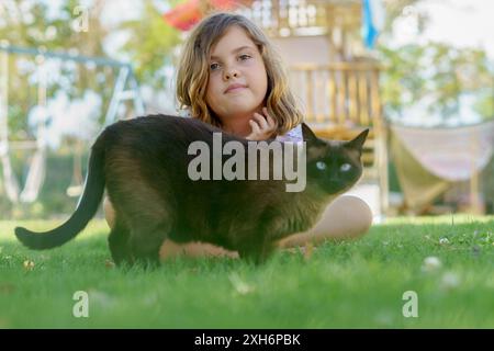 Une jeune fille allongée sur l'herbe avec son chat siamois dans une cour ensoleillée Banque D'Images