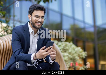 Jeune homme d'affaires assis sur le banc écoutant de la musique avec des écouteurs en utilisant smartphone à l'extérieur. Port du costume et des lunettes, souriant et détendu, immeuble de bureaux en arrière-plan. Banque D'Images