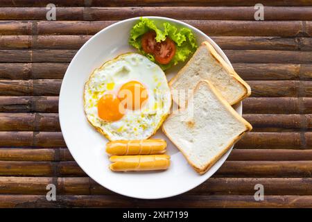 Oeufs brouillés, saucisses, tomates fraîches avec salade verte et toasts sur assiette blanche sur table en bambou brun. Petit déjeuner américain. Vue de dessus. Banque D'Images