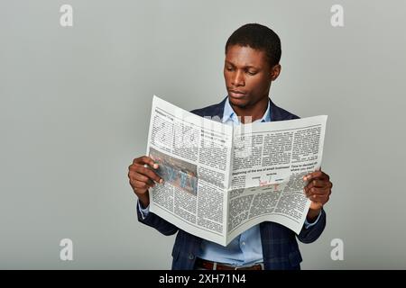 Élégant homme d'affaires afro-américain dans le blazer à carreaux, entièrement immergé dans la lecture d'un journal classique. Banque D'Images