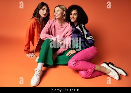 Trois jeunes femmes multiculturelles vêtues de vêtements élégants sont assises par terre, posant pour une photo amusante sur fond orange. Banque D'Images