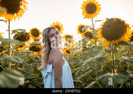 Une jeune femme aux longs cheveux blonds sourit alors qu'elle se tient debout dans un champ de tournesols, illuminée par le soleil couchant. Banque D'Images