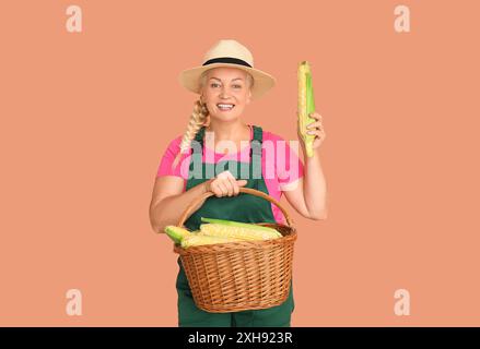 Heureuse fermière mature avec panier en osier plein d'épis de maïs mûrs sur fond orange Banque D'Images