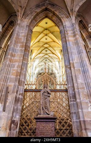 Intérieur de la cathédrale Saint Vitus à Prague, République tchèque. La photo montre une statue en pierre d'un personnage religieux debout sur un piédestal, entouré de treillis dorés complexes. Le plafond voûté orné ci-dessus est visible à travers une série d'arches. Banque D'Images