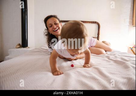 Mère joyeuse et bébé jouent ensemble sur un lit confortable dans une pièce confortable remplie de lumière naturelle. Moment réconfortant de liens familiaux et de rire Banque D'Images