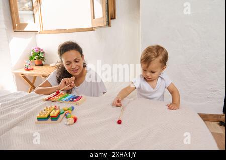 Mère et enfant en bas âge jouant avec des jouets éducatifs colorés sur un lit dans une pièce lumineuse avec la lumière du soleil qui traverse la fenêtre. Banque D'Images