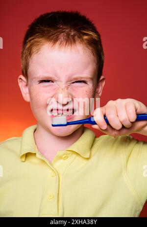 Garçon aux cheveux rouges avec un visage drôle se brosse les dents avec une brosse à dents. Hygiène dentaire. Portrait d'enfant garçon avec brosse à dents isolé sur fond rouge Banque D'Images