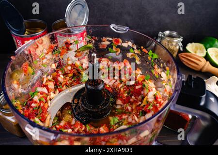 Salsa de blender fraîchement préparée dans un bol de robot culinaire : salsa dans un robot culinaire avec des boîtes de tomates vides, des épices et un citron vert jus en arrière-plan Banque D'Images