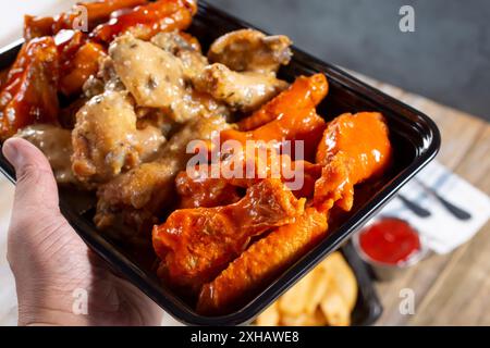 Une vue d'une main tenant un récipient à emporter d'ailes de poulet assorties. Banque D'Images