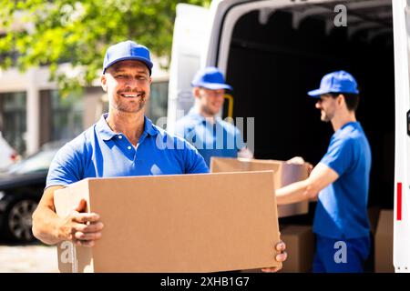 Équipe de livreurs professionnels en uniformes bleus déchargeant efficacement des boîtes en carton d'un camion pour un service de relocalisation majeur. Banque D'Images