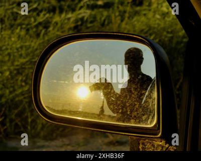 Silhouette du caméraman dans le rétroviseur latéral : la perspective d'une voiture au coucher du soleil. Attraper le soleil. Concept. Banque D'Images