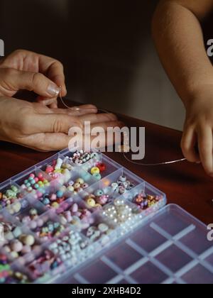Maman et fille créant des bijoux en perles : un gros plan vertical capture deux paires de mains, une adulte et une enfant en bas âge, travaillant ensemble sur un ensemble de perles Banque D'Images