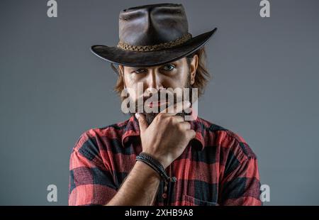 Style cow-boy dans une chemise à carreaux. Chapeau de cow-boy en cuir. Beau jeune homme ajustant son chapeau de cow-boy. Des cowboys en chapeau. Beau macho barbu. Homme non rasé Banque D'Images