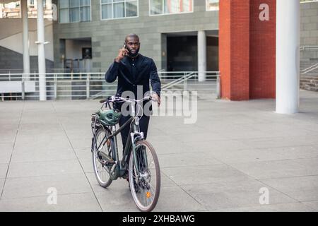 Homme d’affaires africain parlant sur son téléphone portable tout en marchant à côté de son vélo électrique dans le quartier financier de la ville. Il capture un moment de Banque D'Images