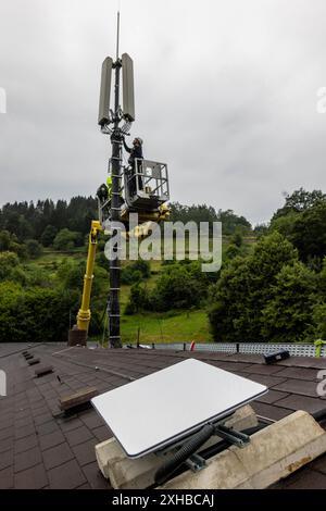 PRODUCTION - 10 juillet 2024, Bade-Württemberg, Forbach : des techniciens se tiennent sur une plate-forme élévatrice et travaillent sur un mât de téléphone portable, tandis qu'une antenne satellite Starlink blanche est visible au premier plan. Vodafone coopère avec la société américaine Starlink dans le cadre d’un projet pilote de couverture de téléphonie mobile dans les zones rurales, dont les satellites peuvent actuellement fournir un accès Internet rapide dans toute l’Europe, l’Amérique du Nord, l’Amérique du Sud et l’Australie. Le fournisseur de téléphonie mobile a installé à cet effet un mât de transmission à Forbach, qui établit le contact avec les serveurs de Vodafone à Francfort via Starlin Banque D'Images