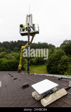 PRODUCTION - 10 juillet 2024, Bade-Württemberg, Forbach : des techniciens se tiennent sur une plate-forme élévatrice et travaillent sur un mât de téléphone portable, tandis qu'une antenne satellite Starlink blanche est visible au premier plan. Vodafone coopère avec la société américaine Starlink dans le cadre d’un projet pilote de couverture de téléphonie mobile dans les zones rurales, dont les satellites peuvent actuellement fournir un accès Internet rapide dans toute l’Europe, l’Amérique du Nord, l’Amérique du Sud et l’Australie. Le fournisseur de téléphonie mobile a installé à cet effet un mât de transmission à Forbach, qui établit le contact avec les serveurs de Vodafone à Francfort via Starlin Banque D'Images