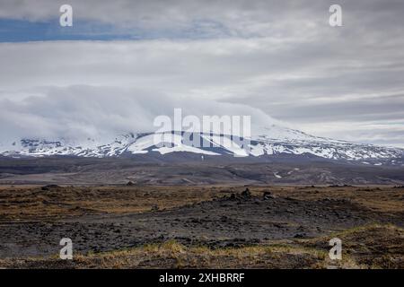 Paysage islandais brut avec champ de lave volcanique et volcan Hekla enneigé avec pic couvert de nuages. Banque D'Images