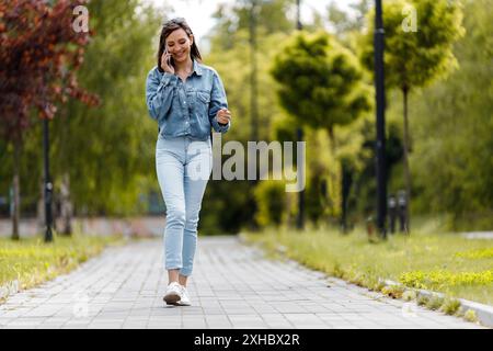 Une femme portant une chemise en denim bleu et un Jean marche le long d'un chemin pavé dans un parc. Elle sourit et parle au téléphone. Banque D'Images