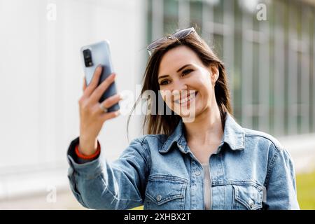 Une femme aux longs cheveux bruns et aux lunettes de soleil prend un selfie avec un smartphone en souriant. Banque D'Images