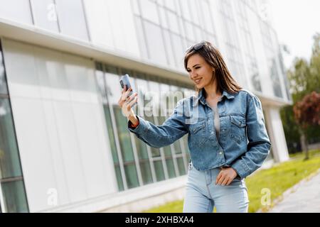 Une femme dans une veste en denim bleu et un Jean prend un selfie avec son téléphone tout en se tenant devant un bâtiment moderne. Banque D'Images