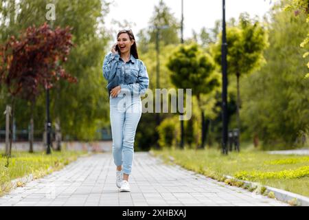 Une femme portant un Jean bleu et une veste en denim marche le long d'un chemin pavé dans un parc, souriant et parlant au téléphone. Banque D'Images