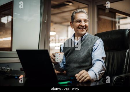 Un homme âgé dans un pull gris et une chemise bleue est assis à un bureau dans un bureau, souriant et tenant une tasse. Il regarde à sa droite, et il y a un lapto Banque D'Images