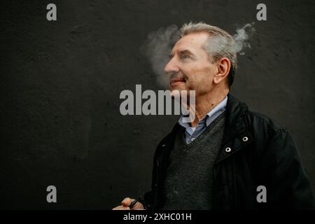 Un homme âgé aux cheveux gris et à la barbe se tient contre un mur gris foncé et expire une bouffée de fumée. Banque D'Images