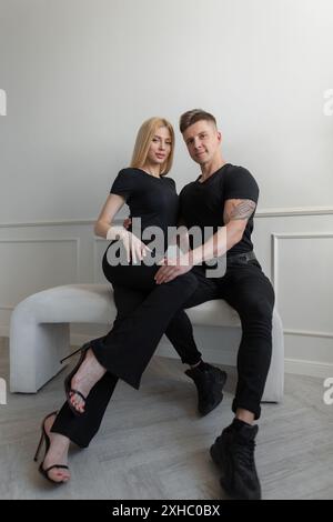 Belle jeune couple de mode femme blonde et homme dans des vêtements noirs à la mode avec un t-shirt assis ensemble près du mur Banque D'Images