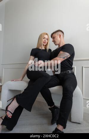 Cool mode beau couple dans des vêtements noirs élégants assis ensemble près d'un mur blanc dans la chambre Banque D'Images
