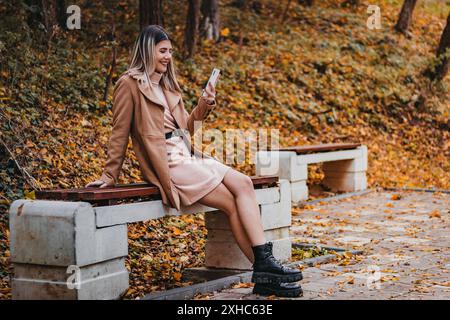 Une femme dans un manteau marron et une robe rose est assise sur un banc de parc à l'automne, regardant son téléphone. Elle est souriante et semble profiter de son temps ou Banque D'Images