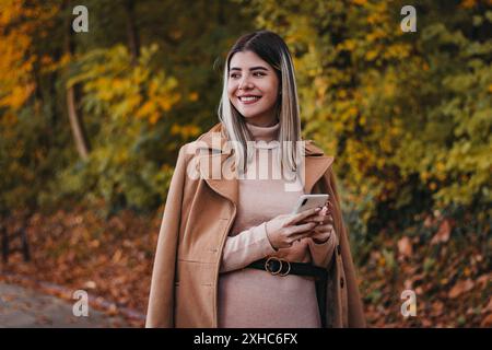 Une femme au manteau brun se tient dans un parc d'automne, souriante et regardant sur le côté tout en tenant un smartphone dans ses mains. Banque D'Images