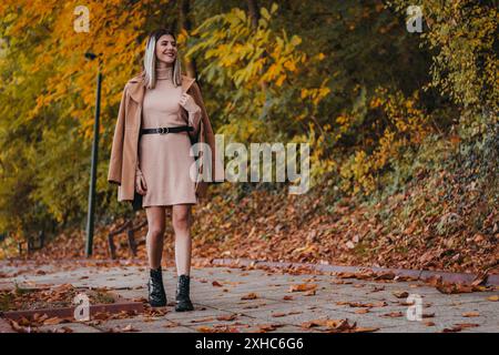 Une femme en robe rose et manteau brun marche sur un chemin entouré de feuilles tombées dans un cadre automnal. Banque D'Images