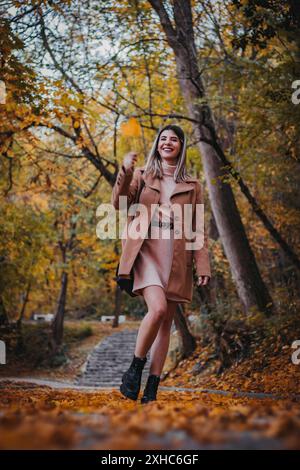Une femme au manteau brun marche à travers une forêt d'automne, souriant alors qu'elle tient une feuille tombée. Banque D'Images
