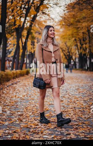 Une femme traverse un chemin couvert de feuilles d'automne, vêtue d'un manteau brun et de bottes noires. Banque D'Images