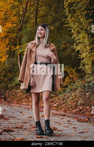 Une femme dans une robe rose et un manteau brun marche dans un chemin couvert de feuilles d'automne. Banque D'Images