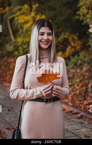 Une femme dans un pull rose tient une feuille orange sèche dans un parc avec des feuilles mortes sur le sol. Banque D'Images