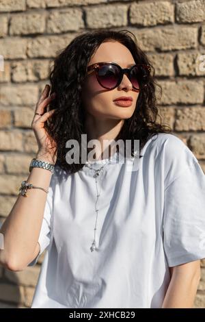 Belle fille d'été élégante avec coiffure bouclée avec des lunettes de soleil dans un T-shirt blanc à la mode près d'un mur de briques dans la rue Banque D'Images