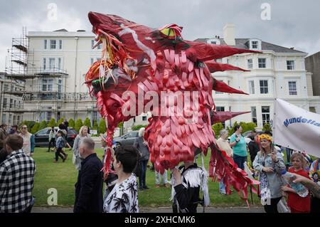 Folkestone, Kent, Royaume-Uni. 13 juillet 2024. Défilé annuel du Charivari Day. Folklore britannique et traditions rituelles sont le thème de la saisissante parade du carnaval de cette année. Des sculptures massives de tête ornent les épaules des jeunes locaux avec des enfants de l'école primaire à travers le district habillant aux couleurs du thème de l'année. Marchant du stade, en passant par la ville jusqu’au kiosque à musique Leas, le Charivari Day reste la plus grande tradition coutumière de Folkestone depuis 1997 et attire des centaines de spectateurs dans la ville portuaire en bord de mer. Crédit : Guy Corbishley/Alamy Live News Banque D'Images