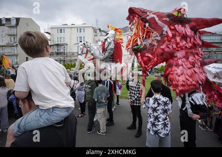 Folkestone, Kent, Royaume-Uni. 13 juillet 2024. Défilé annuel du Charivari Day. Folklore britannique et traditions rituelles sont le thème de la saisissante parade du carnaval de cette année. Des sculptures massives de tête ornent les épaules des jeunes locaux avec des enfants de l'école primaire à travers le district habillant aux couleurs du thème de l'année. Marchant du stade, en passant par la ville jusqu’au kiosque à musique Leas, le Charivari Day reste la plus grande tradition coutumière de Folkestone depuis 1997 et attire des centaines de spectateurs dans la ville portuaire en bord de mer. Crédit : Guy Corbishley/Alamy Live News Banque D'Images