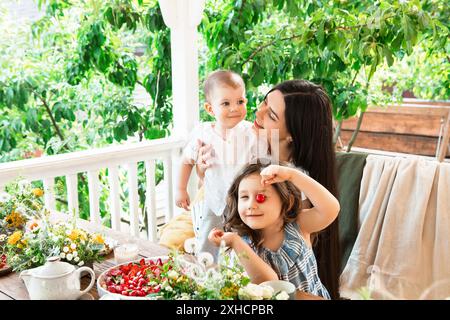 Femme souriante assise sur le banc avec des frères et sœurs adorables à une table en bois pendant le petit déjeuner dans l'arrière-cour Banque D'Images