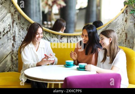 Une jeune femme joyeuse partage des données sur son smartphone avec diverses amies tout en étant assise autour d'une table et en dégustant un café dans un café confortable Banque D'Images