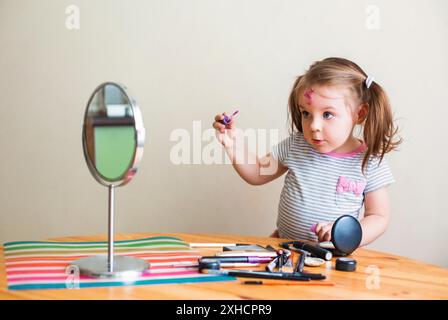 Adorable petit enfant avec le visage sale jouant avec des cosmétiques et faisant le maquillage tout en regardant dans le miroir Banque D'Images