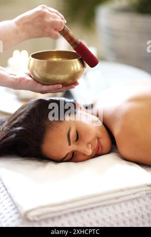 Vue latérale d'une femme thérapeute utilisant un bol de chant au-dessus de l'arrière de la femme asiatique détendue dans le salon de spa Banque D'Images