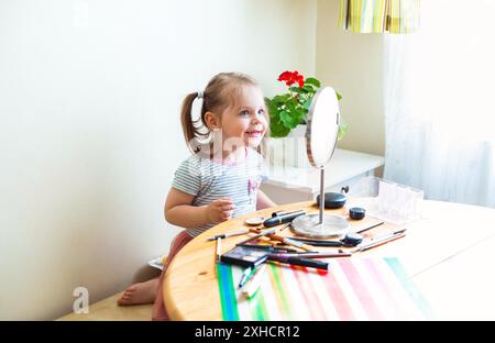 Adorable petit enfant avec le visage sale jouant avec des cosmétiques et faisant le maquillage tout en regardant dans le miroir Banque D'Images