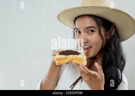 Vue rapprochée de la belle jeune femme asiatique en chapeau de paille et montrant du pain blanc avec des saupoudrages de chocolat souriant doucement, isolé sur blanc, breakfa Banque D'Images