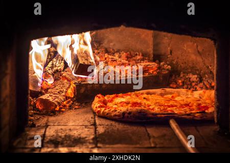 Une seule pizza cuit dans un four en briques avec un feu rugissant et des braises incandescentes. Banque D'Images