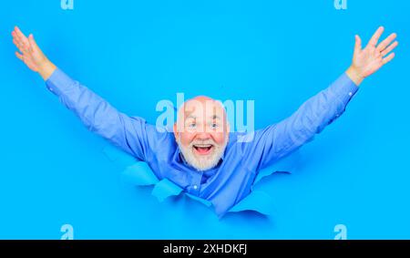 Heureux homme barbu regarde à travers le trou dans le papier bleu. Vieil homme souriant dans une chemise élégante avec les mains levées. Casser le papier. Espace de copie pour le texte ou le slogan. Banque D'Images